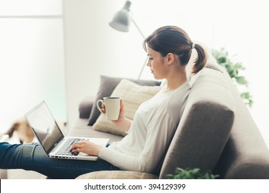 Young Woman Relaxing At Home On The Couch, She Is Having A Coffee And Using A Laptop