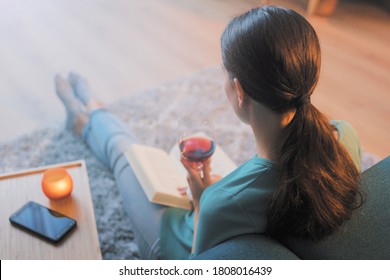 Young Woman Relaxing At Home: She Is Sitting On The Floor In The Living Room, Drinking A Glass Of Wine And Reading A Book