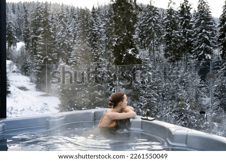 Young woman relaxing in a hot tub and thermal spa enjoying a winter view of snowy Christmas trees. Concept of recreation on hot bath and escape to nature