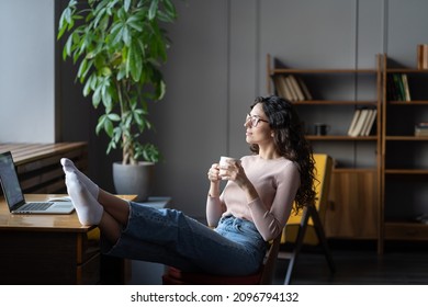 Young woman relaxing at office or cozy home workplace with bare feet on desk drinking tea or coffee. Happy satisfied millennial female freelancer rest sitting at laptop think of future success concept - Shutterstock ID 2096794132
