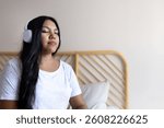 Young woman relaxing on bed, listening to music with wireless headphones, finding peace and tranquility in her bedroom