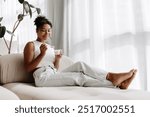 Young woman is relaxing on her couch at home, enjoying a cup of yogurt and smiling