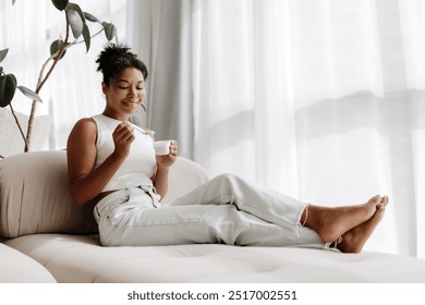 Young woman is relaxing on her couch at home, enjoying a cup of yogurt and smiling - Powered by Shutterstock