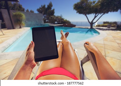 Young woman relaxing on a lounge chair using a tablet PC near the pool. User POV. Female model sitting on a deckchair holding digital tablet. - Powered by Shutterstock