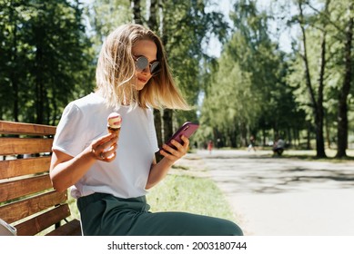 Young woman relaxing on a park bench and using a mobile phone. Nice girl eating ice cream and chatting with someone on a smartphone, outdoors. Hipster girl vacation summer lifestyle, side view - Powered by Shutterstock