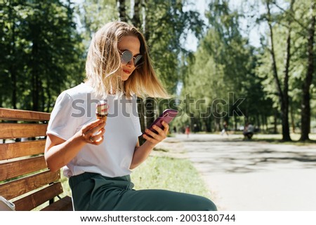 Young woman relaxing on a park bench and using a mobile phone. Nice girl eating ice cream and chatting with someone on a smartphone, outdoors. Hipster girl vacation summer lifestyle, side view