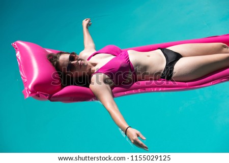 Young woman relaxing on a pink raft in an outdoor swimming pool in a bikini and aviator sunglasses looking at the camera. Enjoying the sun, lounging in a swimming pool arms extended.