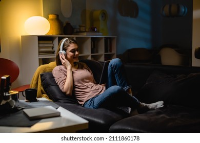 Young Woman Relaxing On Sofa At Home. She Is Using Digital Tablet And Listening Music On Headphones . Night Evening Scene