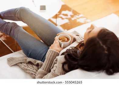 Young woman relaxing on sofa holding cup with lemon tea. Woman dreaming in living room lying on couch with legs on table and looking up. Woman thinking while drinking lemon tea at home.