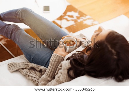 Young woman relaxing on sofa holding cup with lemon tea. Woman dreaming in living room lying on couch with legs on table and looking up. Woman thinking while drinking lemon tea at home.
