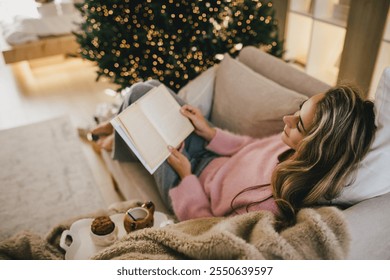 Young woman relaxing, reading book and drinking cocoa in a cozy festive living room near Christmas tree. - Powered by Shutterstock