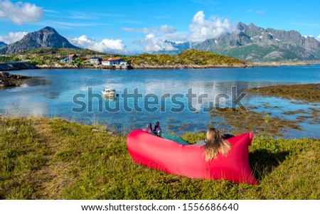 Young woman is relaxing in the red airlounger in the border of Vestfjord of Lofoten archipelago. Mountain landscape of Austvagoya island are at background. Nordland, Northern Norway.

