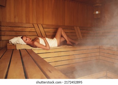 Young woman relaxing and sweating in hot sauna wrapped in towel. Girl In Sauna. Interior of Finnish sauna, classic wooden sauna with hot steam. Russian bathroom. Relax in hot bathhouse with steam.
