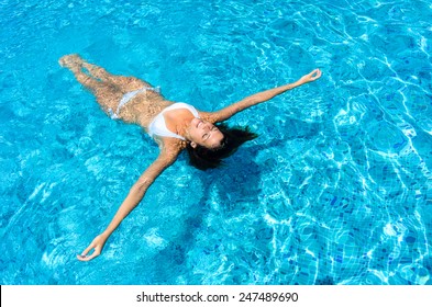 Young woman relaxing in swimming pool on summer vacation. Brunette caucasian model in white bikini floating in water.