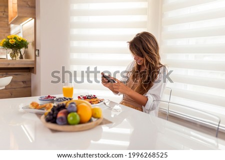 Young woman relaxing while having breakfast at home, photographing food using smart phone