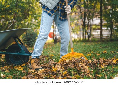 A young woman removes fallen autumn leaves with a rake in the garden of a country house.