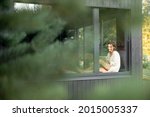 Young woman resting at beautiful country house or hotel, sitting with tablet on the window sill enjoying beautiful view on pine forest. View from outside. Beautiful destinations for vacation