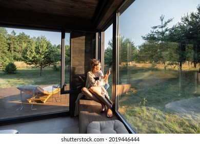 Young Woman Resting At Beautiful Country House Or Hotel, Sitting On The Window Sill Enjoying Beautiful View On Pine Forest And Making A Photo On Phone. Concept Of Solitude And Recreation On Nature