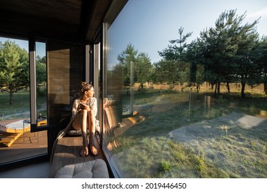 Junge Frau, die sich in einem schönen Landhaus oder Hotel, sitzend auf dem Fenster ruht, genießen einen schönen Blick auf Kiefernwald. Konzept der Einsamkeit und Erholung in der Natur