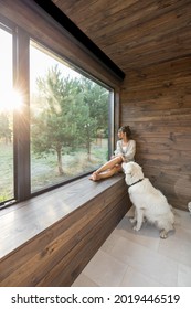 Young Woman Resting At Beautiful Country House Or Hotel, Sitting On The Window Sill With Pine Forest View While Sunrise And White Dog Sits Near. Concept Of Solitude And Recreation On Nature With Pet