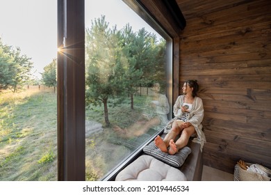 Young Woman Resting At Beautiful Country House Or Hotel, Sitting With Phone And Cup On The Window Sill Enjoying Beautiful View On Pine Forest. Concept Of Solitude And Recreation On Nature