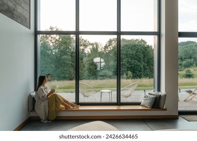 Young woman resting at beautiful country house or hotel, sitting on the window sill enjoying beautiful view on pine forest. Concept of solitude and recreation on nature rest  - Powered by Shutterstock