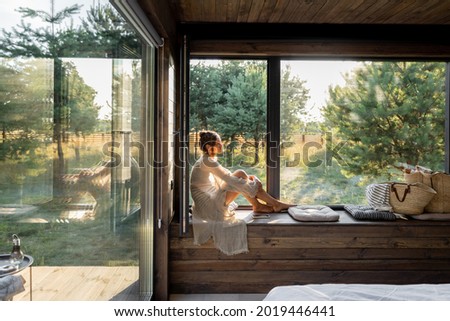 Young woman resting at beautiful country house or hotel, sitting on the window sill enjoying beautiful view on pine forest. Concept of solitude and recreation on nature