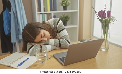 A young woman resting head on desk beside laptop and flowers in a well-lit home office - Powered by Shutterstock