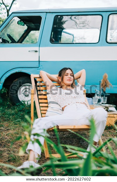 Young woman\
resting in nature, sitting on wooden chair near van  outdoors.\
Enjoying summer, travel\
concept