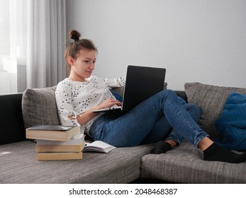 A young woman is resting on the couch and using a laptop. Books and notebooks are lying next to her. The student learns at home. E-learning.