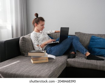 A young woman is resting on the couch and using a laptop. Books and notebooks are lying next to her. The student learns at home. E-learning.