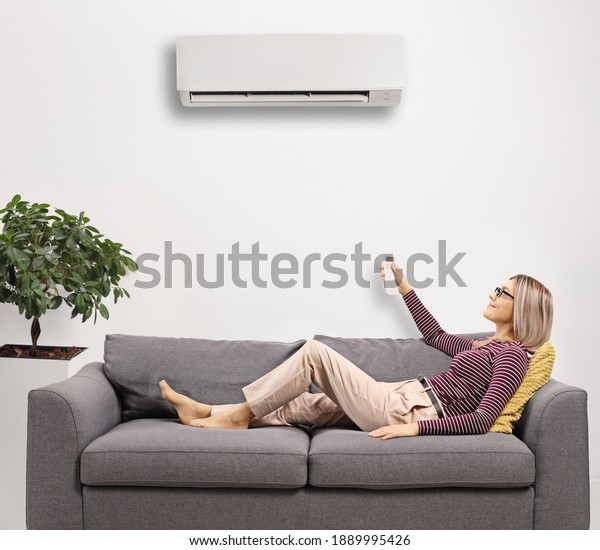 Young woman resting on a gray sofa and\
holding a remote control from an ac unit at\
home