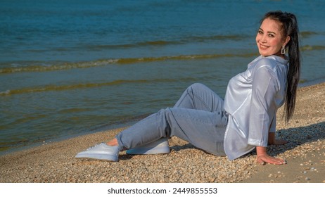 A young woman is resting on the seashore and smiling broadly at her happiness. Dressed in a white shirt, denim trousers and sneakers. - Powered by Shutterstock