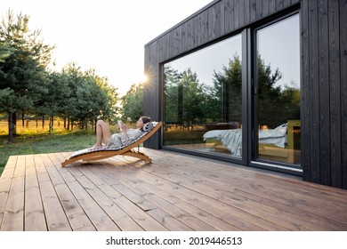 Junge Frau, die sich auf dem Sonnenbett ausruht und auf einem Tablet auf der Holzterrasse neben dem modernen Haus mit Panoramafenstern in der Nähe des Kiefernwaldes liest. Konzept der Einsamkeit und Erholung in der Natur