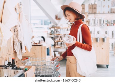 Young woman with reusable cotton bag and empty jar doing shopping in plastic free store. Minimalist vegan style girl buying groceries without plastic packaging in zero waste shop. Low waste lifestyle.