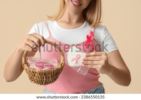 Young woman with ribbon and bagged cookies on beige background, closeup. Breast cancer awareness concept
