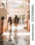 Young woman rider in equestrian uniform in stable saddles horse against background of bright rays of light. Horse riding training. She throws saddle on back of horse. Authentic village barn.