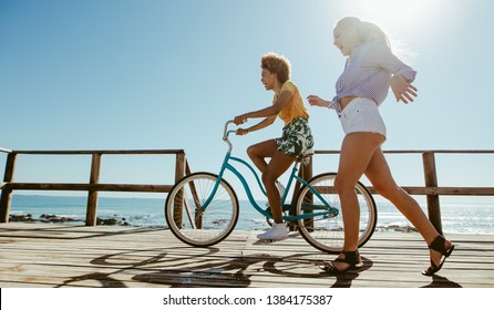 Young Woman Riding A Bicycle With Her Friend Running By On Boardwalk. Multi-ethnic Female Friends Having Fun With A Bike At The Seaside Boardwalk.