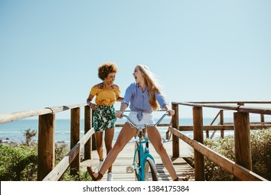 Young Woman Riding Bike On Boardwalk With Friend Running By. Multi-ethnic Girls Having Fun With A Bike At The Seaside Boardwalk.