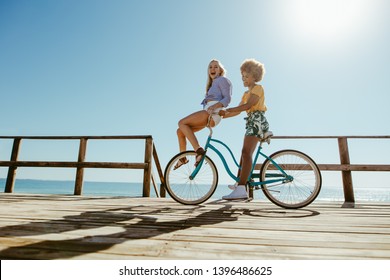 Young woman riding a cycle with her friend sitting on the handlebar on seaside boardwalk. Two girls on enjoying bike ride on a summer day. - Powered by Shutterstock