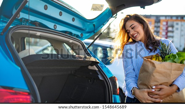 Young woman riding shopping cart full of food on
the outdoor parking. Young woman in car park, loading shopping into
boot of car. Shopping successfully done. Woman putting bags into
car after shopping