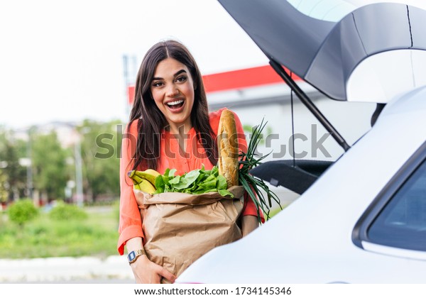 Young woman riding shopping cart full of food on\
the outdoor parking. Young woman in car park, loading shopping into\
boot of car. Shopping successfully done. Woman putting bags into\
car after shopping