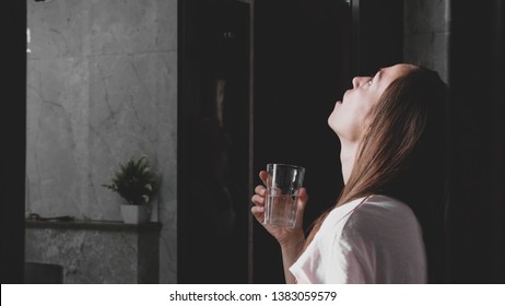 Young woman rinses her mouth with water while looking at mirror and spits water into the sink, side view.