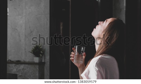 Young woman rinses her\
mouth with water while looking at mirror and spits water into the\
sink, side view.