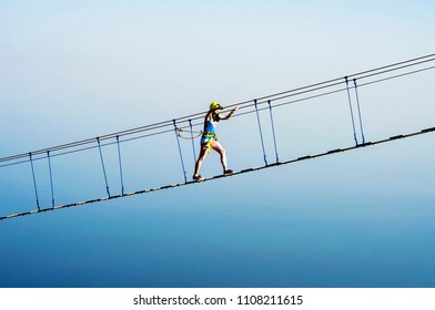 Young Woman Rising On Hanging Rope Bridge At Sea And Sky Background. Leisure Extreme Activity At Summer Day