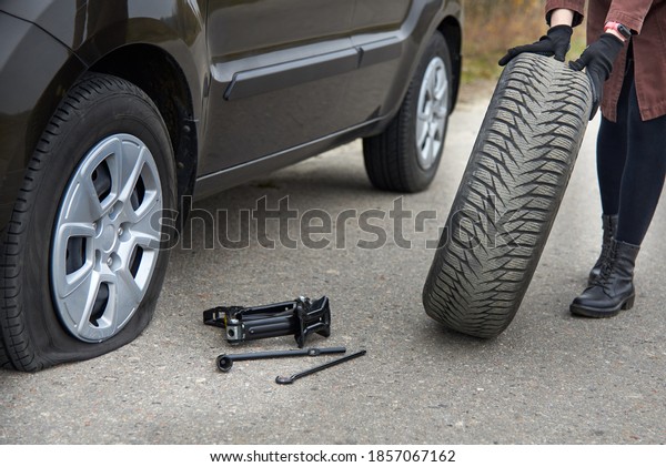 A young woman rolls spare tire near her car\
with a flat tire, trouble on the\
road.