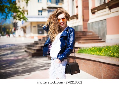Young woman in round hipster sunglasses and leather jacket with long curly hair smiling posing in an urban context. Stylish purse, sity street.