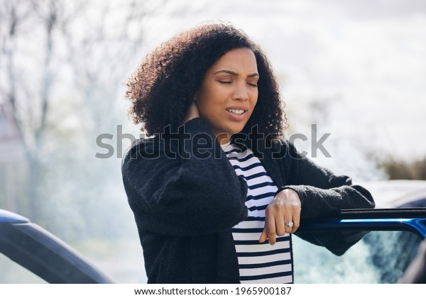 Young woman rubbing neck in\
pain from whiplash injury standing by damaged car after traffic\
accident