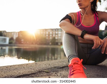 Young woman runner resting after workout session on sunny morning. Female fitness model sitting on street along pond in city. Female jogger taking a break from running workout. - Shutterstock ID 314302445