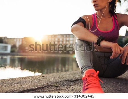 Young woman runner resting after workout session on sunny morning. Female fitness model sitting on street along pond in city. Female jogger taking a break from running workout.